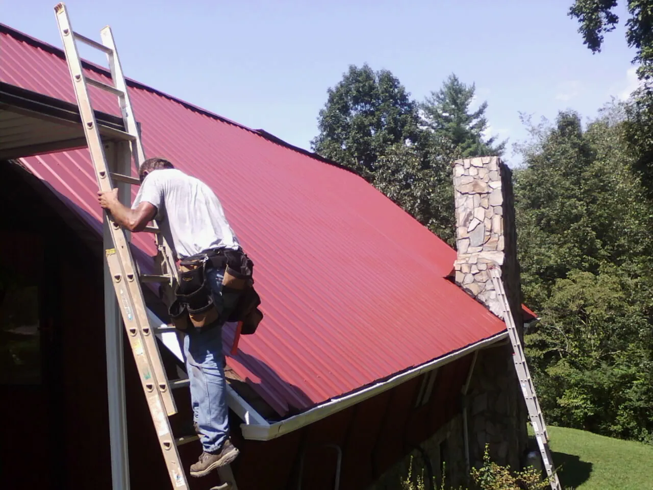Roofer on a ladder beside a red metal roof and stone chimney — Soffit & Fascia Repair by Roofing Brothers in Saint Matthews